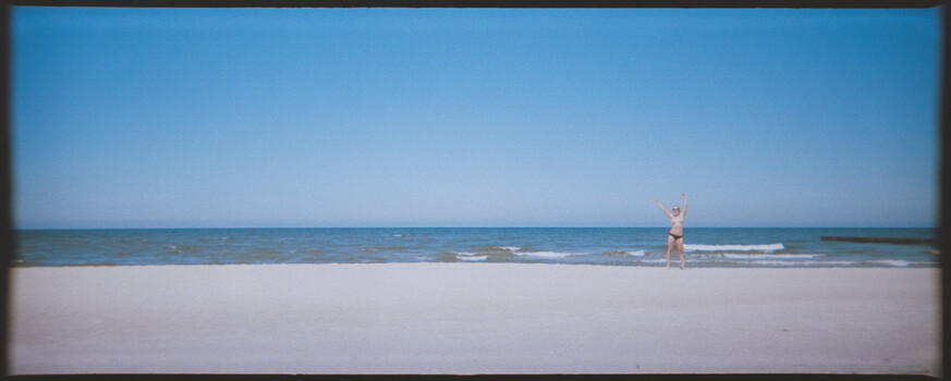 Lone figure standing in shallow ocean water on a wide, empty beach under clear blue sky.