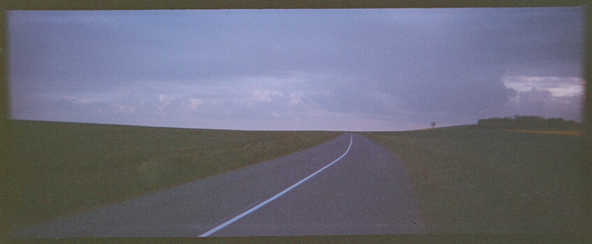 Empty rural road stretching toward the horizon with flat fields on either side under a stormy sky.