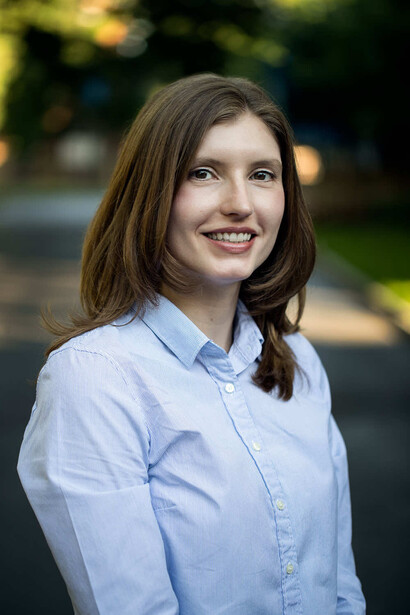 Woman in light blue button-up shirt smiling outdoors with blurred green background.